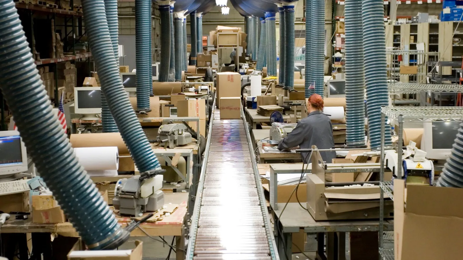 Factory conveyor belt running through workspace with machinery and workers handling packages in a large warehouse.