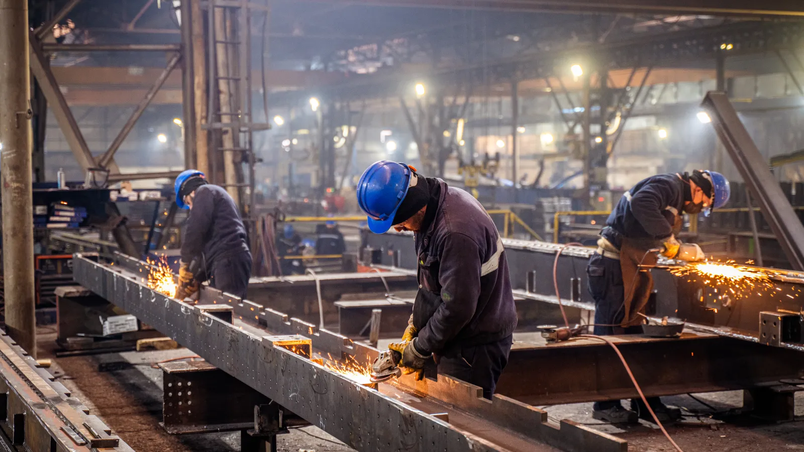 Three workers wearing blue helmets grinding large metal beams in an industrial factory with sparks flying.
