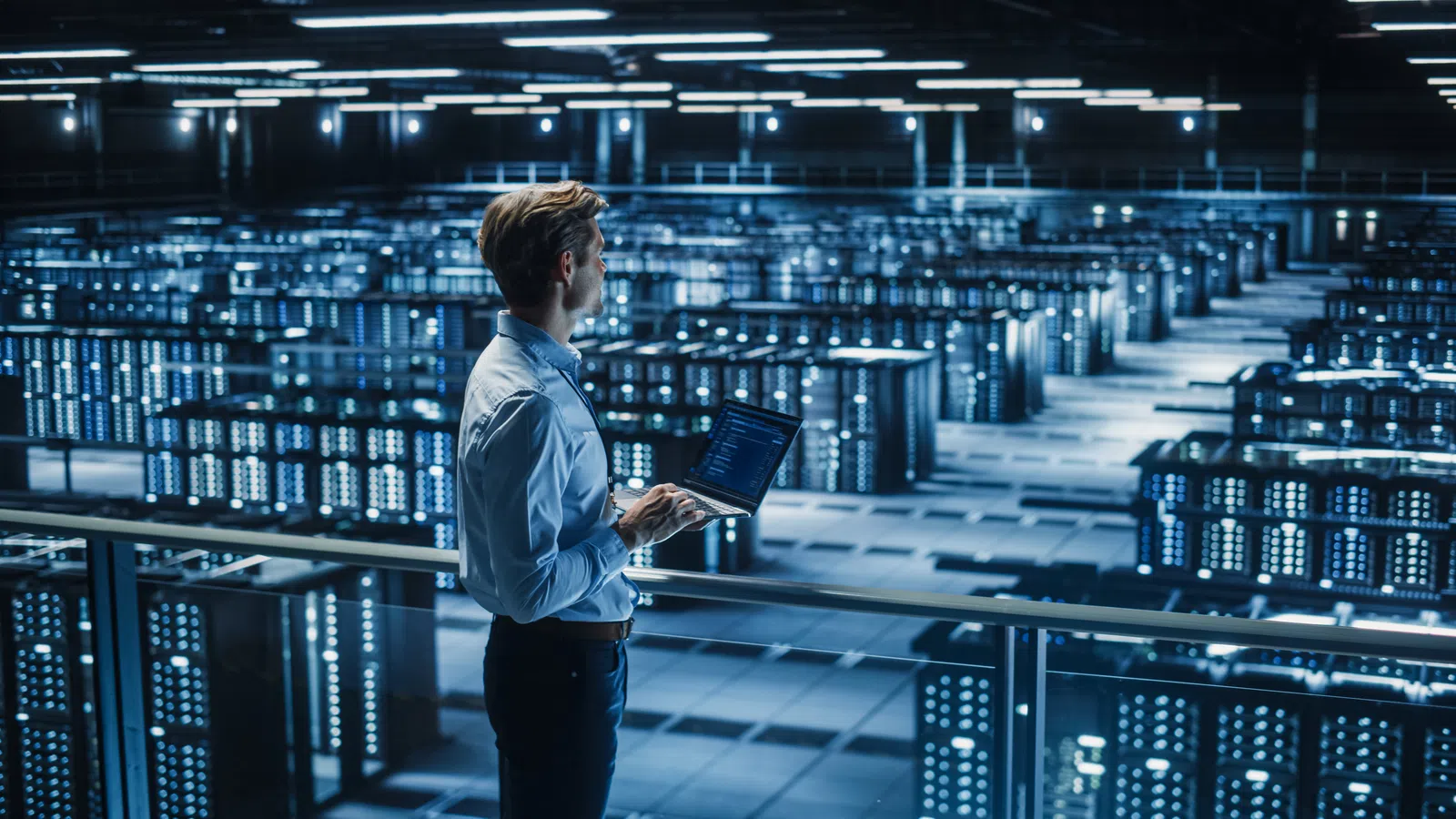 Technician with laptop monitoring server racks in a large data center with blue LED lights.