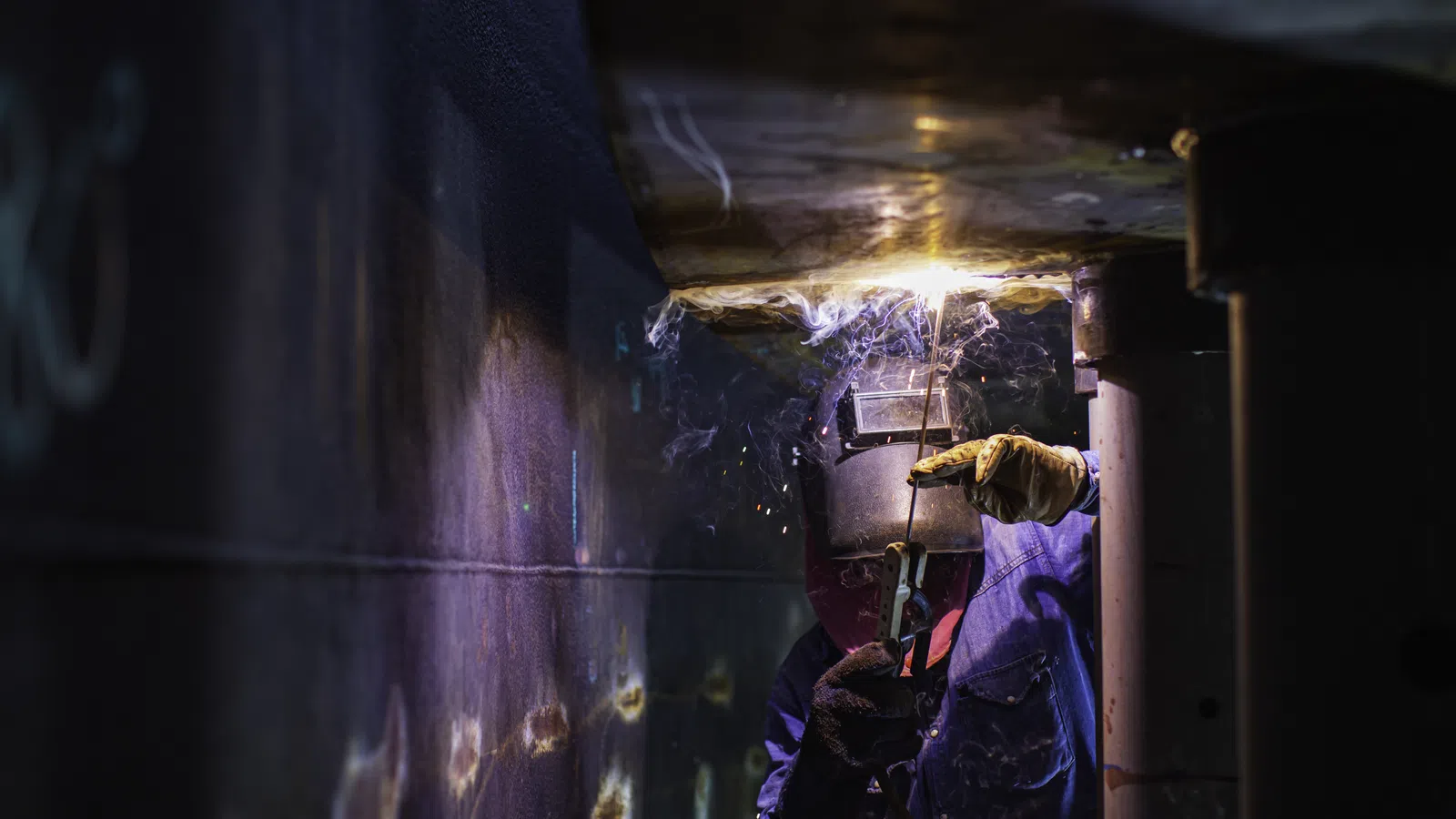 Welder wearing protective gear welding metal with sparks and smoke in a dark industrial setting.