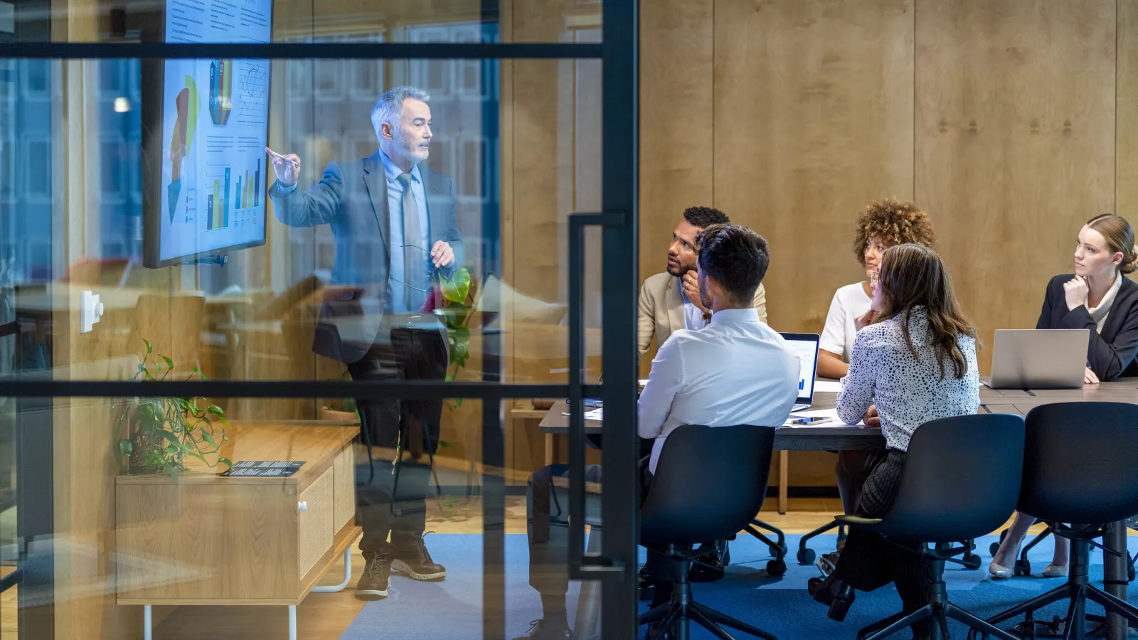 Business team in formal attire attending a presentation with charts in a modern glass office meeting room.