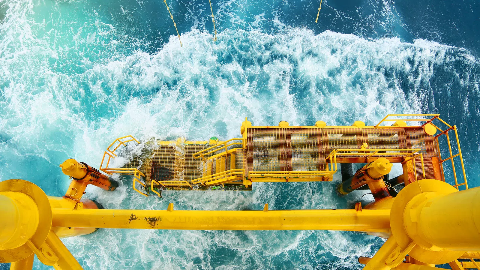 Top-down view of a yellow offshore platform structure above vibrant blue ocean waves and white foam.