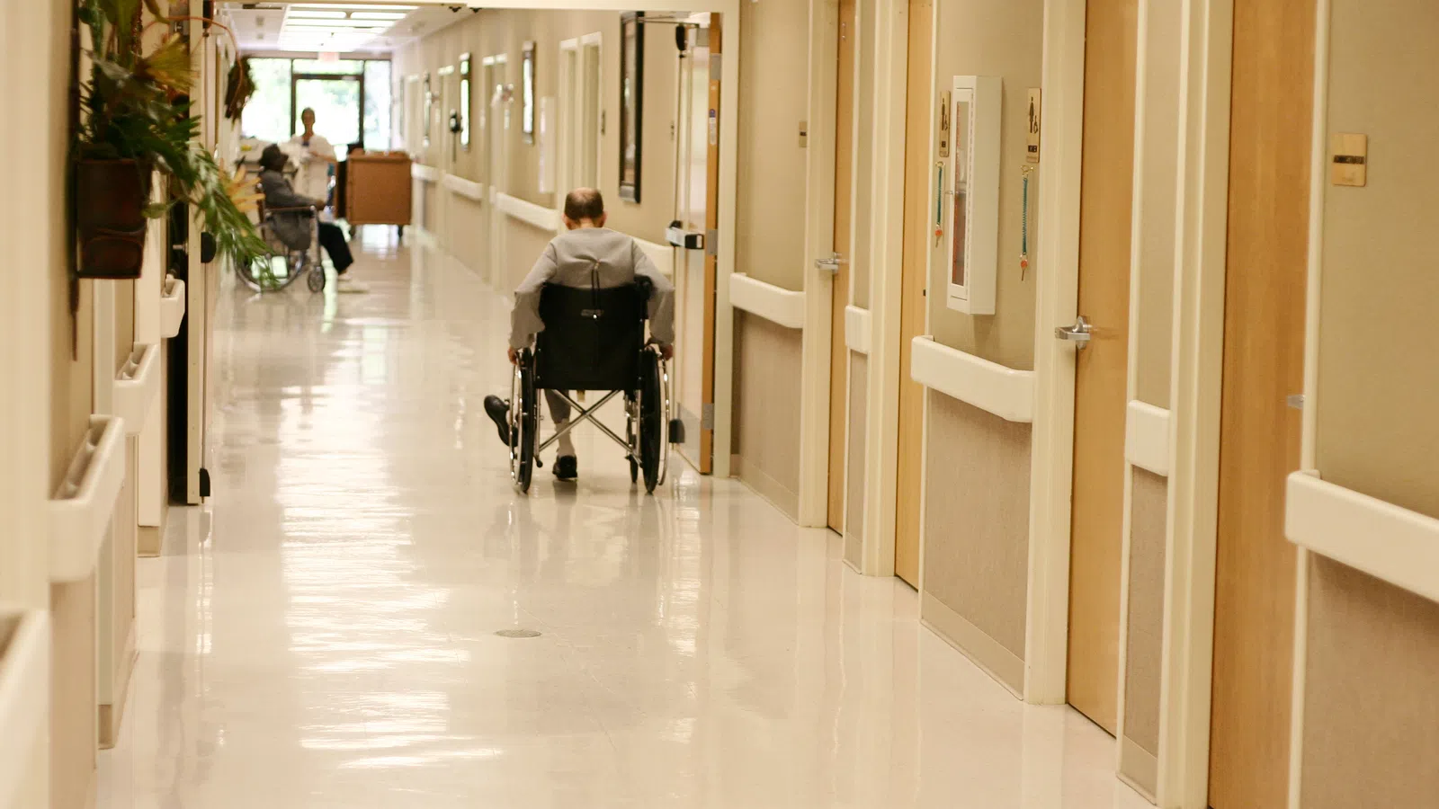 Man in wheelchair moving down a clean, well-lit hospital corridor with multiple closed doors visible.