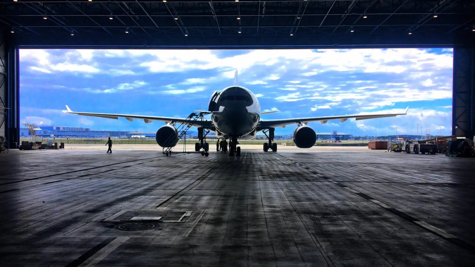 Commercial airplane parked inside a large hangar with maintenance crew and bright blue sky outside.