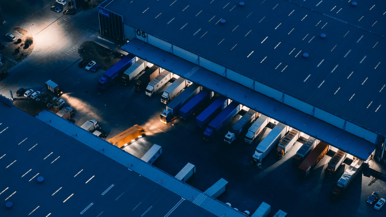 Aerial view of a warehouse loading dock with semi trucks parked and some headlights turned on at dusk.
