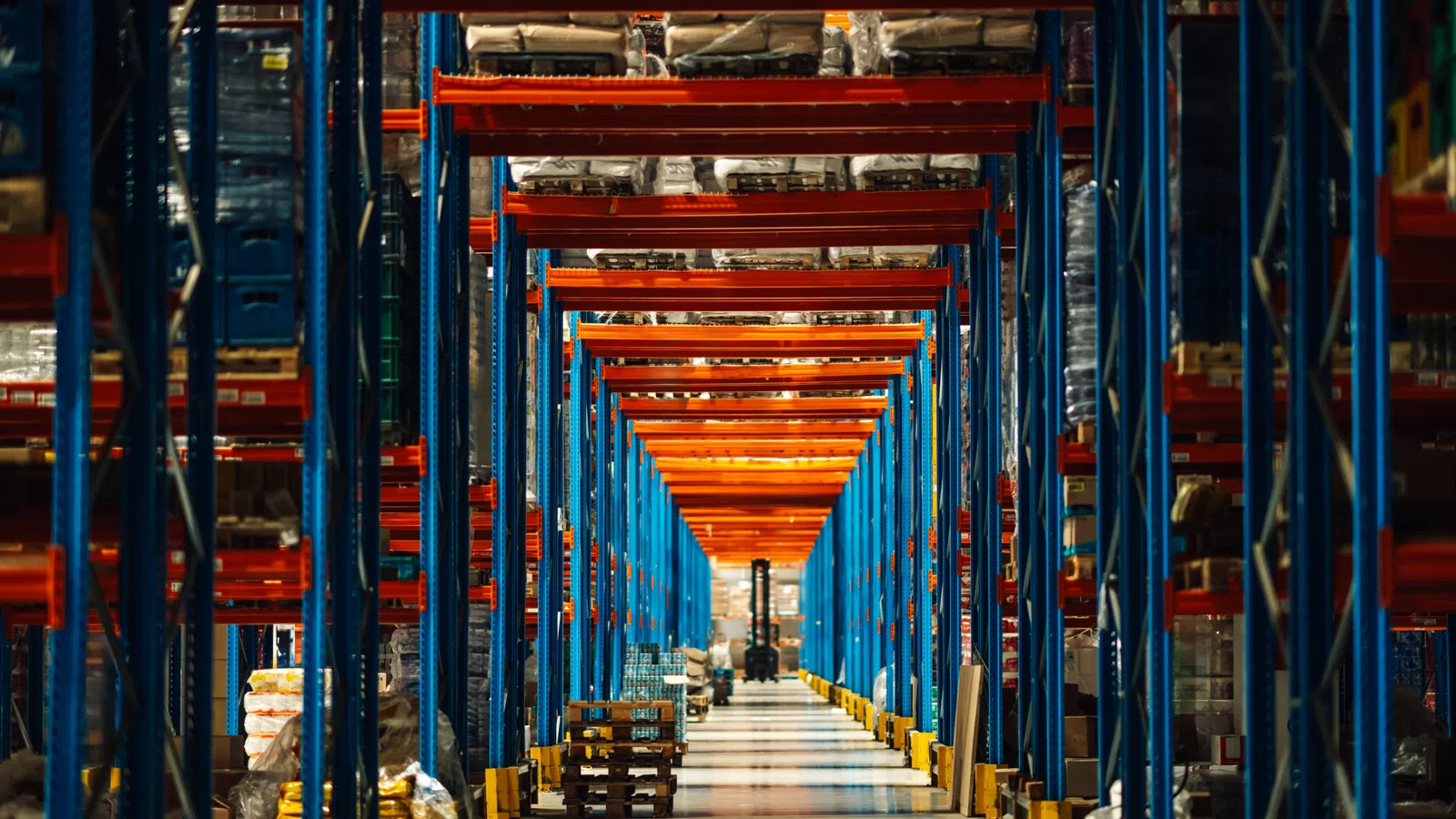 Long aisle view inside a warehouse with blue and orange industrial shelving holding various packaged goods.