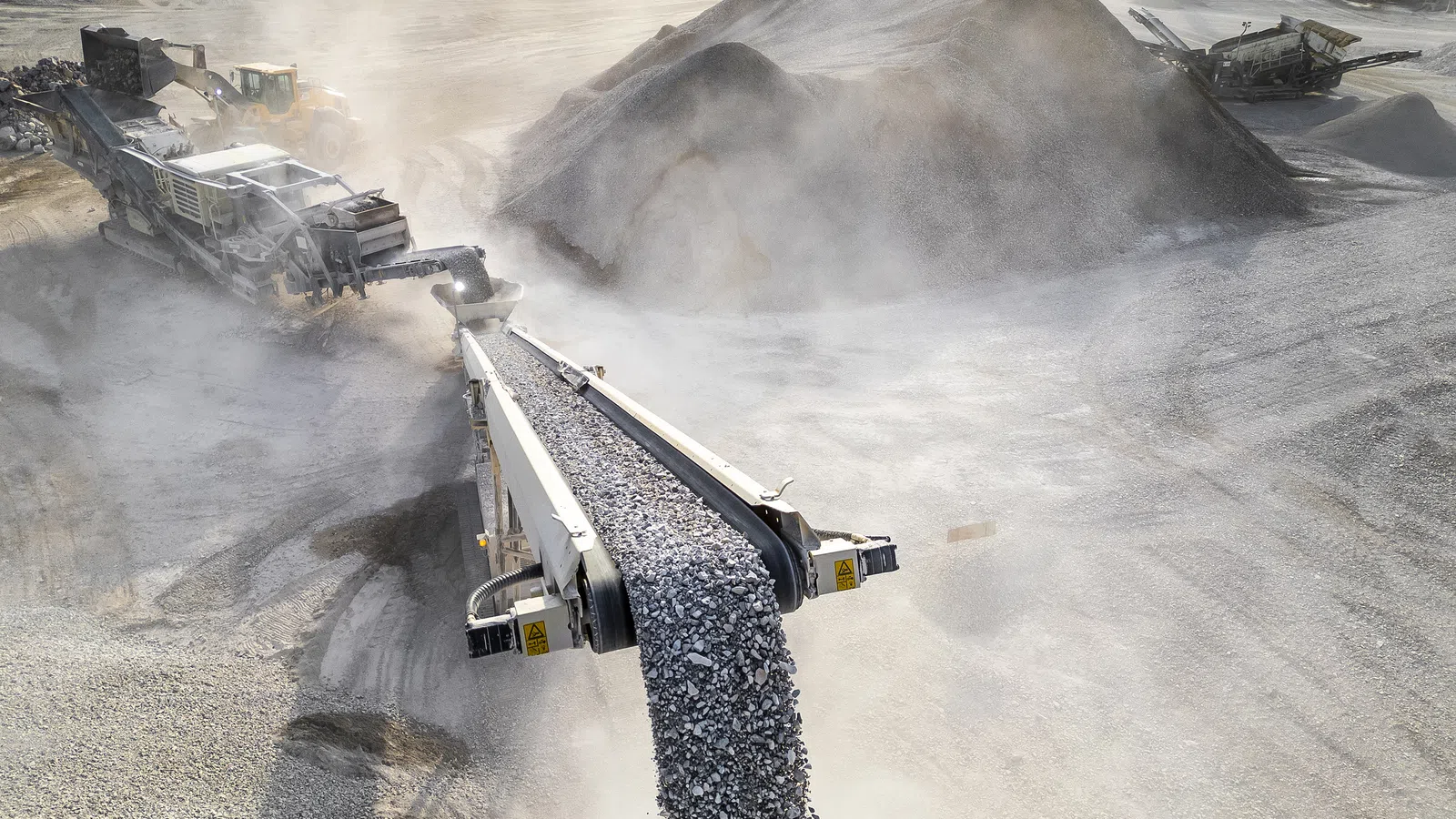 Industrial conveyor belt transporting gravel in a dusty open quarry with heavy machinery and large aggregate piles.