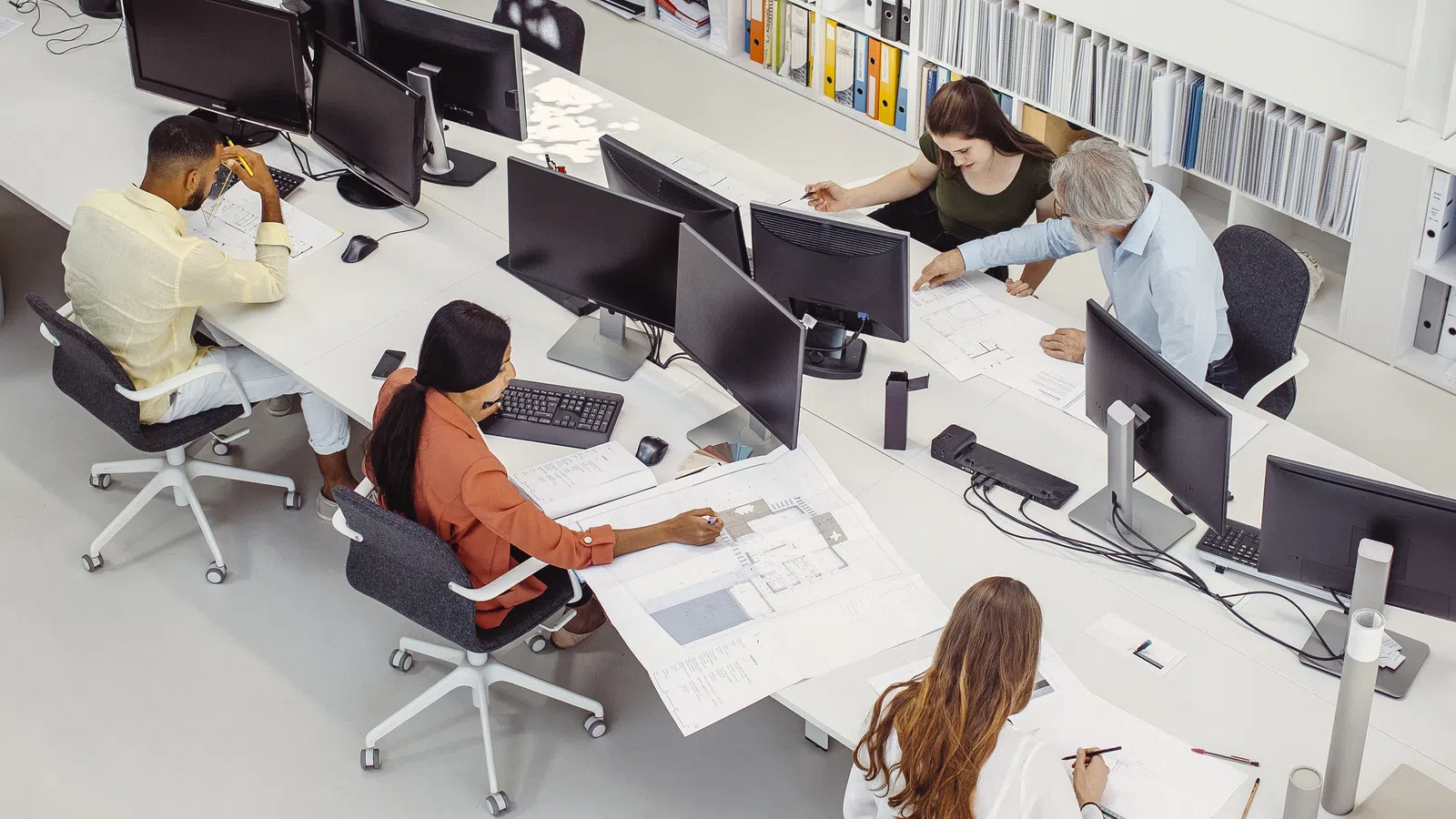 Overhead view of a diverse office team collaborating on architectural plans around multiple computer monitors.