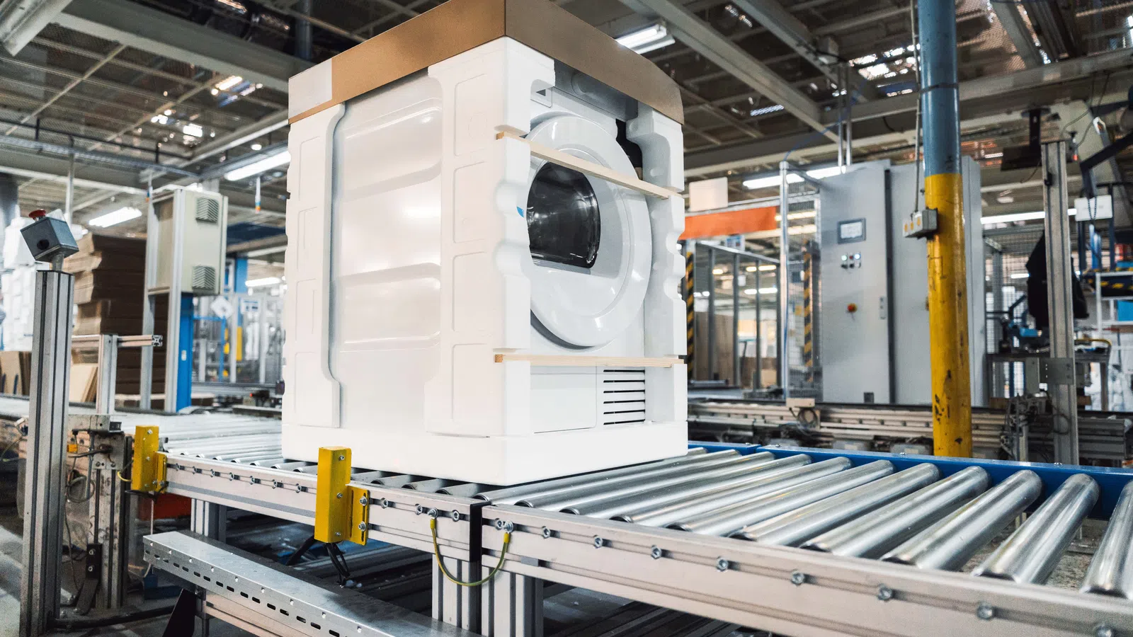 Washing machine packaged in foam on conveyor belt inside a large industrial warehouse factory.