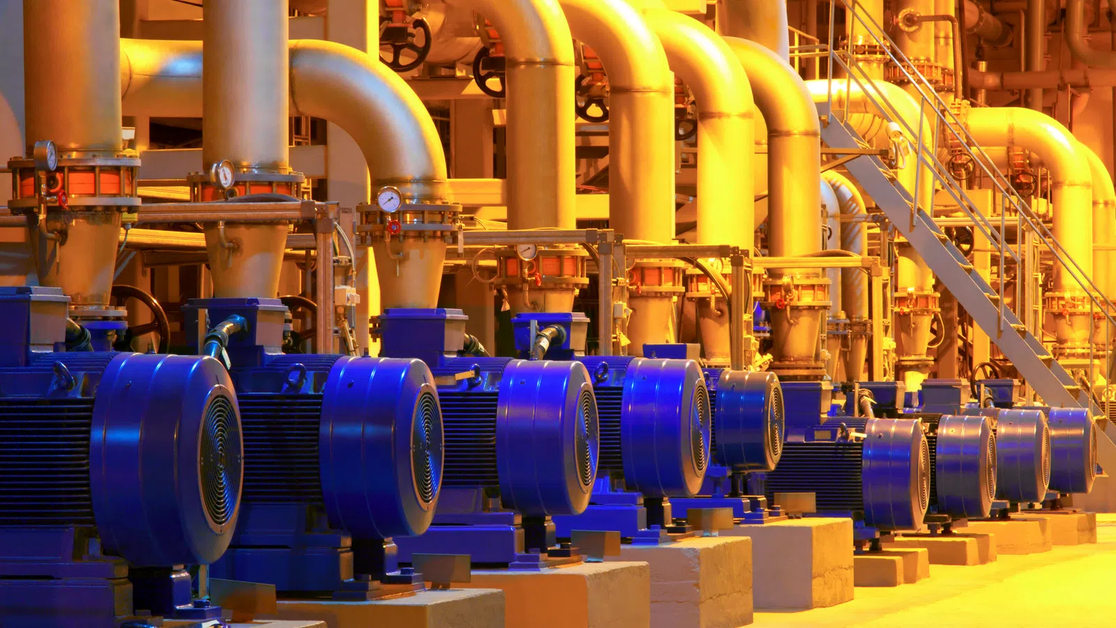Row of blue industrial pumps with large white pipes and staircase in a brightly lit factory setting