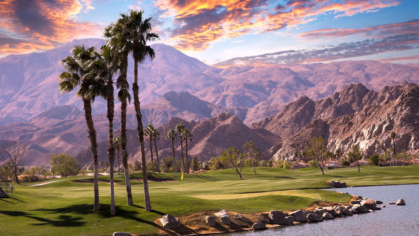 Palm trees and a golf course by a lake with mountains and a colorful sky at sunset.