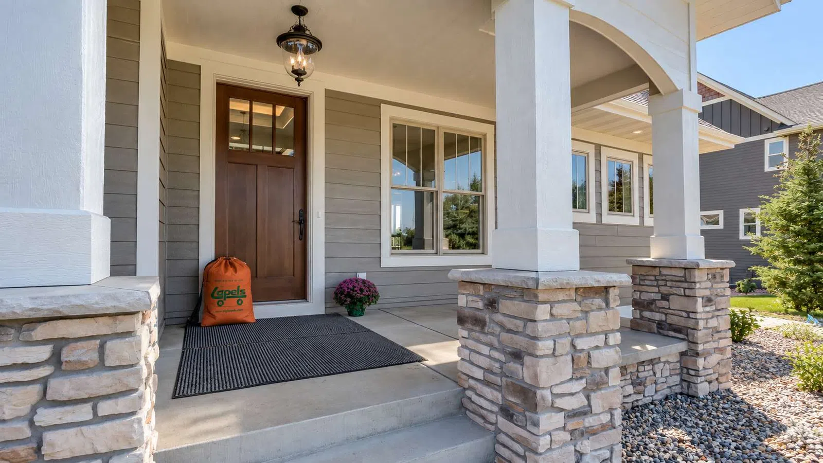 Front porch with stone pillars, wooden door, welcome mat, and decorative potted flowers in a suburban home.