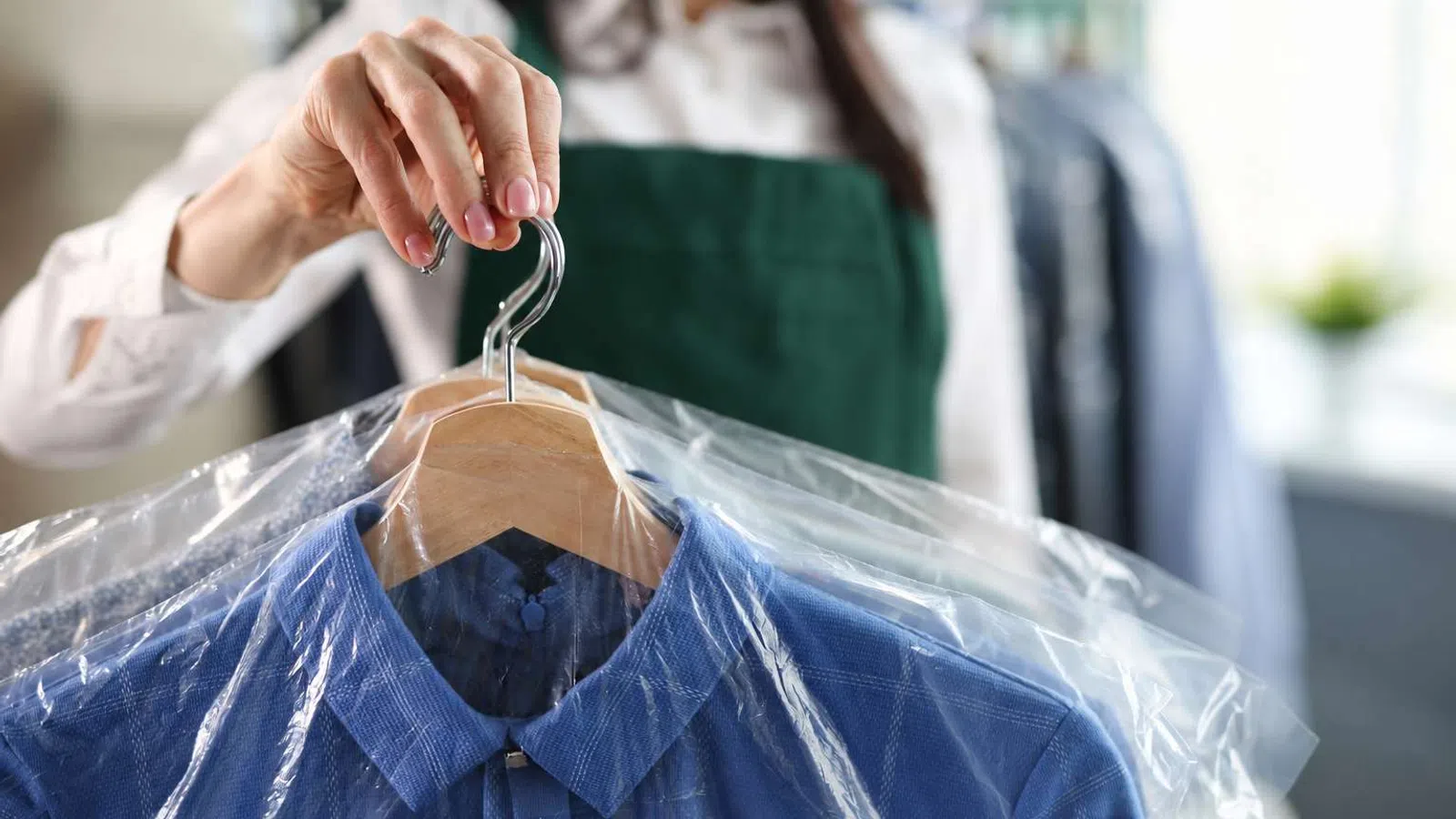 Person holding freshly dry-cleaned blue shirts on wooden hangers covered in plastic garment bags.