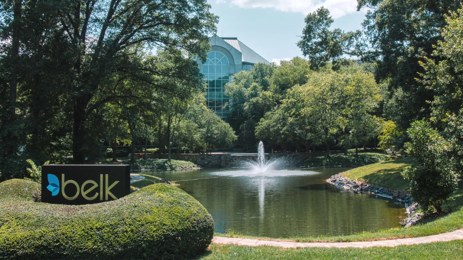Belk company sign by pond with fountain surrounded by lush green trees on a sunny day.