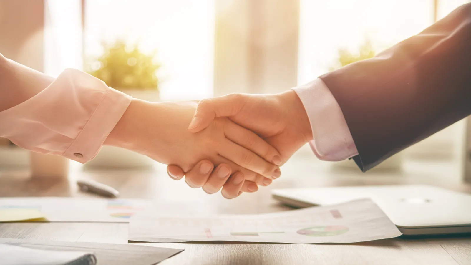 Two professionals shaking hands over a desk with business documents in a bright office setting.