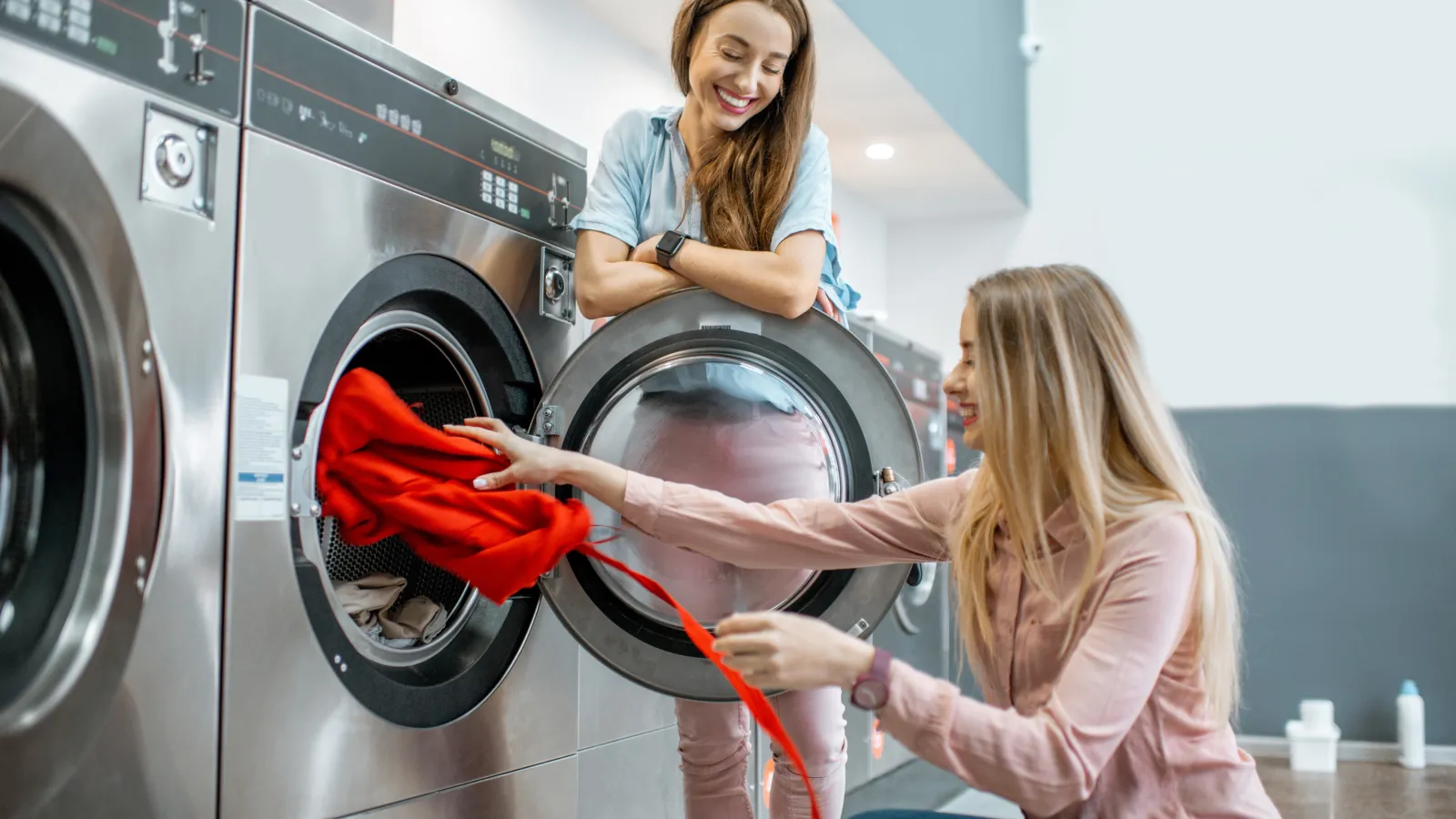 Two women smiling while doing laundry together at a laundromat with stainless steel machines.
