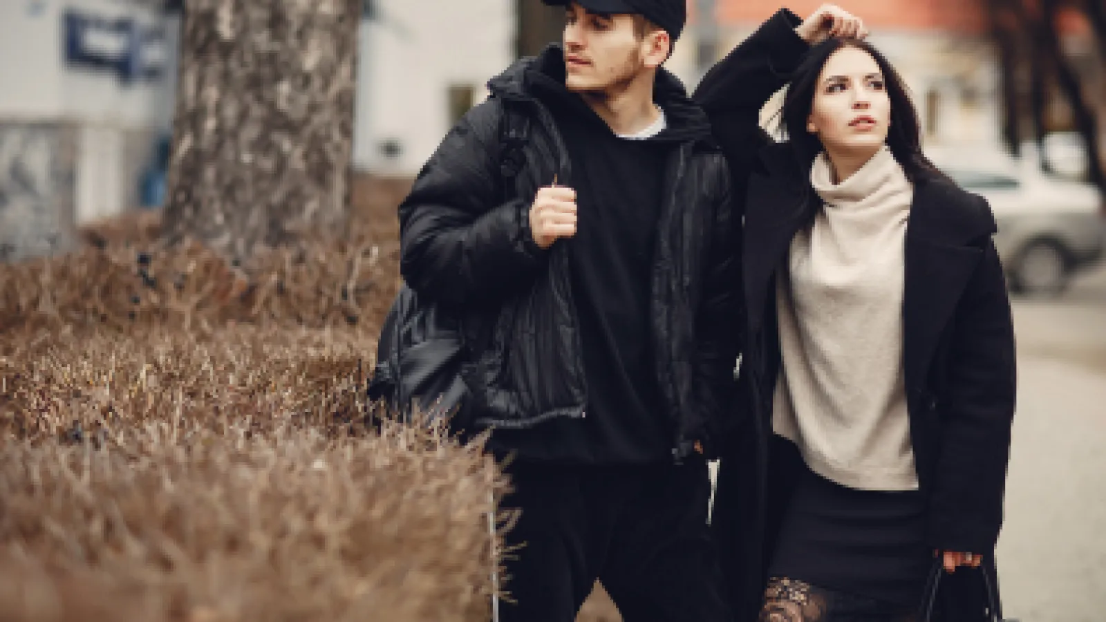 Young man and woman wearing black coats standing outdoors near dried bushes in autumn city setting.