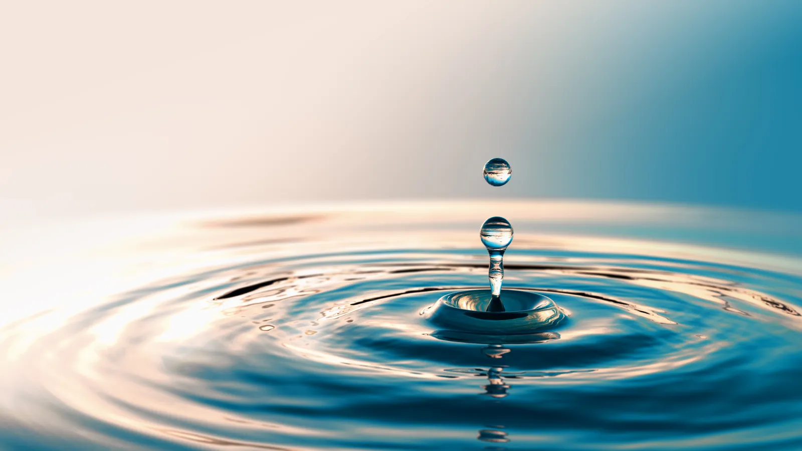 Close-up of a water droplet suspended above rippling water surface with gradient blue and beige background