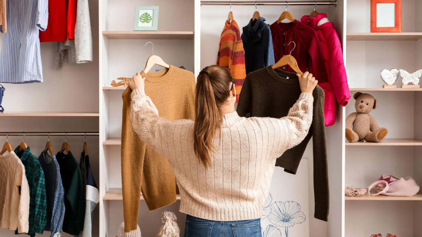 Woman choosing between two sweaters in a well-organized wardrobe with clothes and toys.