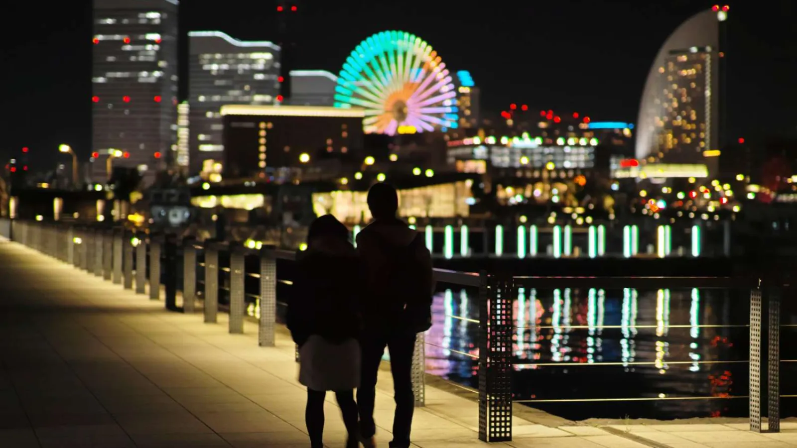 Couple walking on illuminated waterfront promenade at night with city skyline and colorful Ferris wheel in background