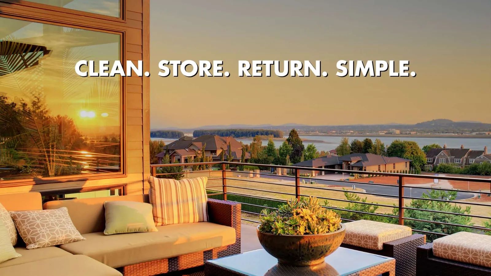 Sunset view from a cozy outdoor patio with cushioned seating, plants, and distant houses by the water.