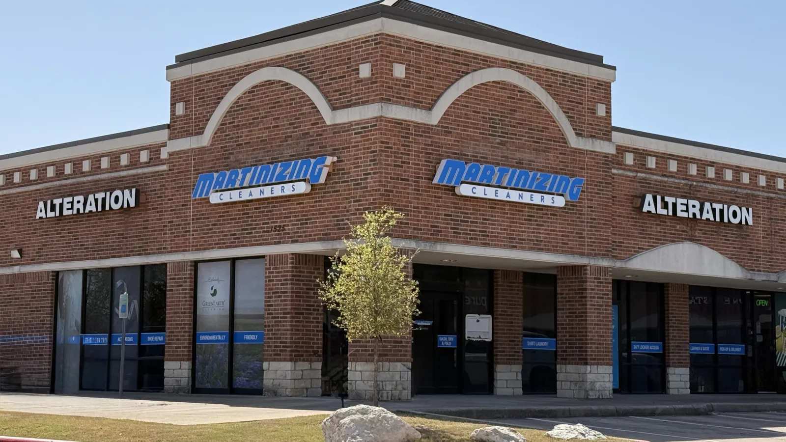 Brick corner storefront of Martinizing Cleaners with alteration signs under clear blue sky and small tree in front
