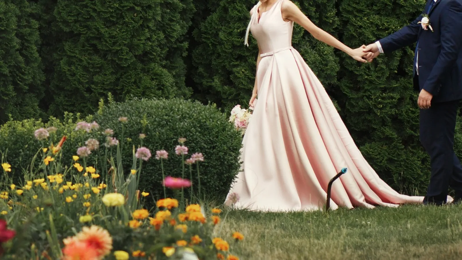 Bride in a pink gown and groom in navy suit holding hands in garden with colorful flowers and green bushes.