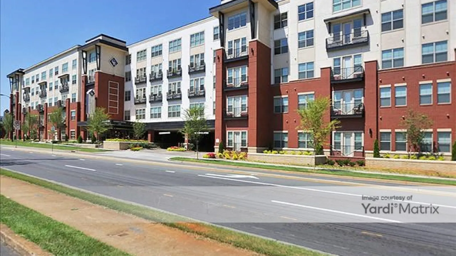 Modern multi-story apartment building with red and white facade along a wide street under clear blue sky.