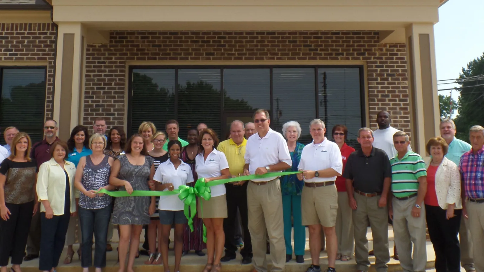 Group of people at a ribbon-cutting ceremony outside Lapels Dry Cleaning store with bright sunny weather.