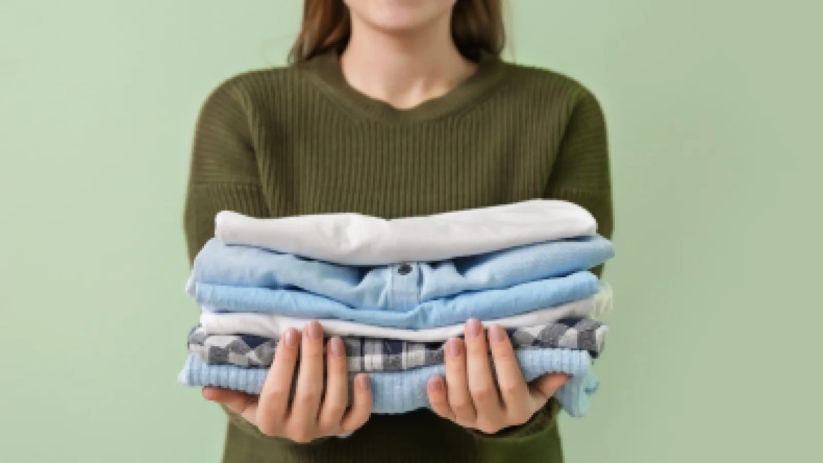 Smiling woman in green sweater holding a stack of neatly folded blue and white clothes against a green background
