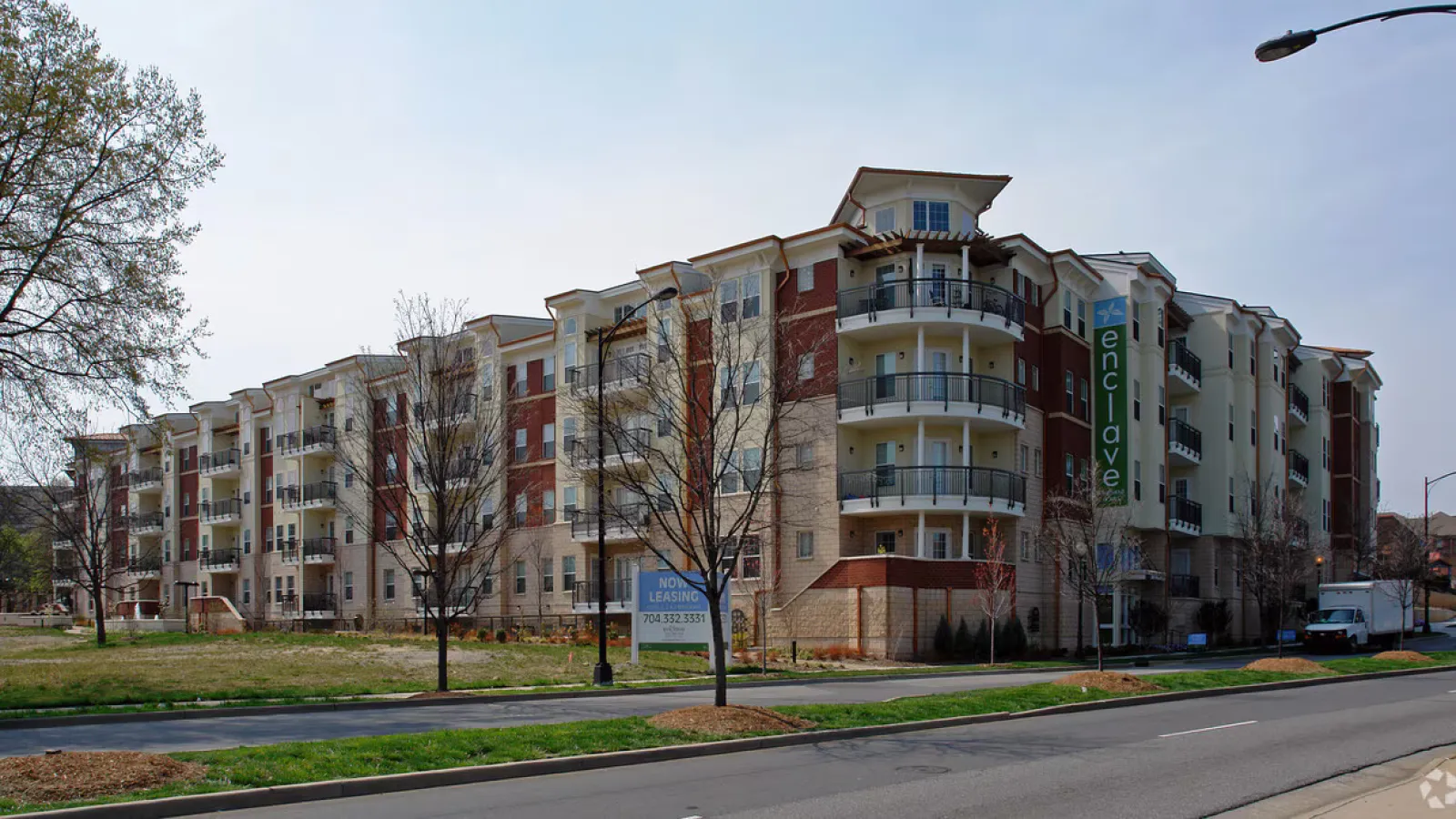 Modern multi-story apartment building with balconies and trees along a quiet street under a clear sky