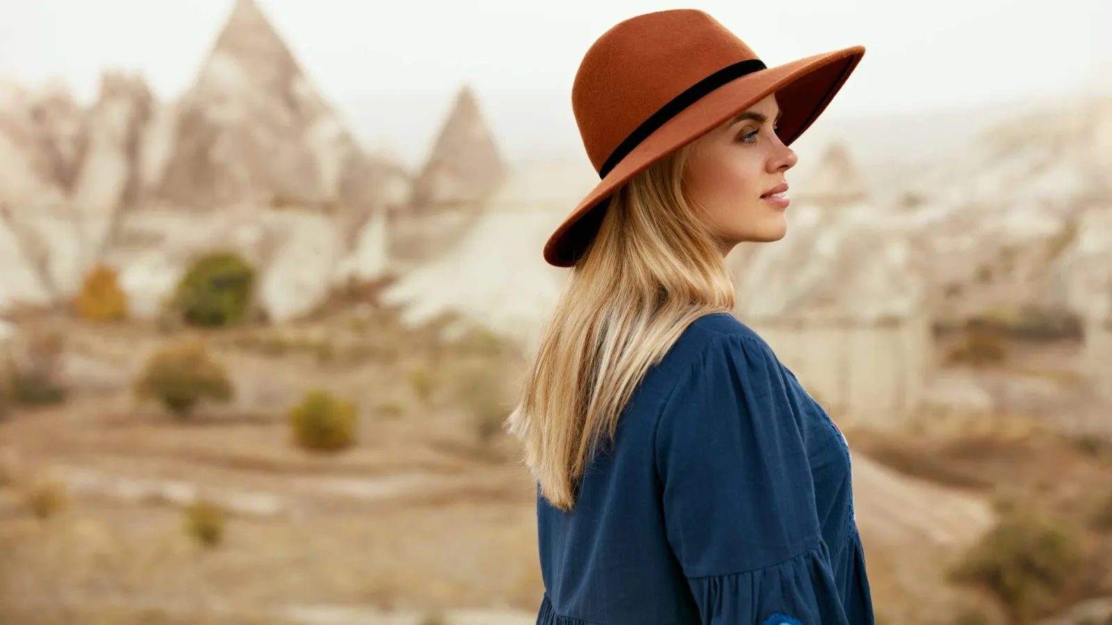 Woman wearing a brown hat and blue dress stands outdoors with rocky hills in the background under soft light.