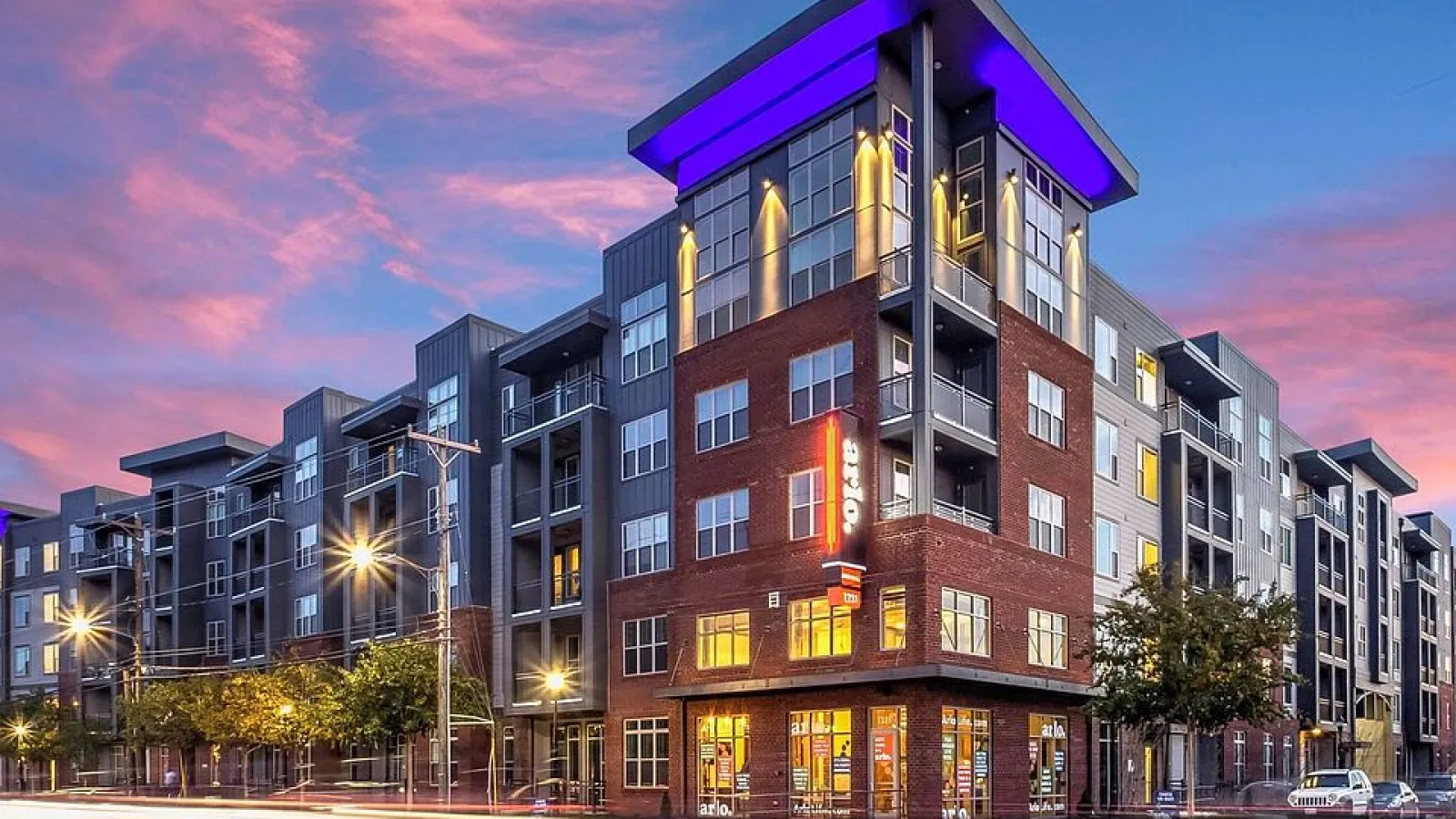 Modern multi-story apartment building with illuminated storefronts and purple rooftop lighting at sunset.