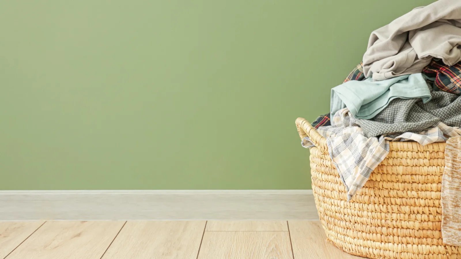 Woven laundry basket overflowing with clothes on wooden floor against green wall background