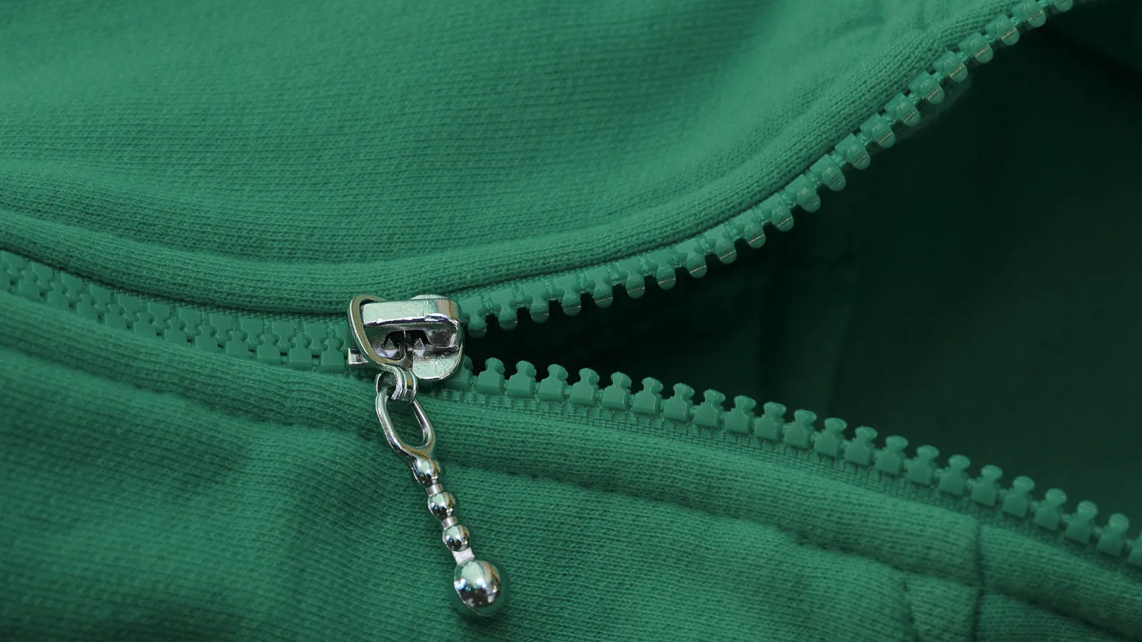 Close-up of a green zipper with a metal pull on textured fabric, partially unzipped showing dark interior.