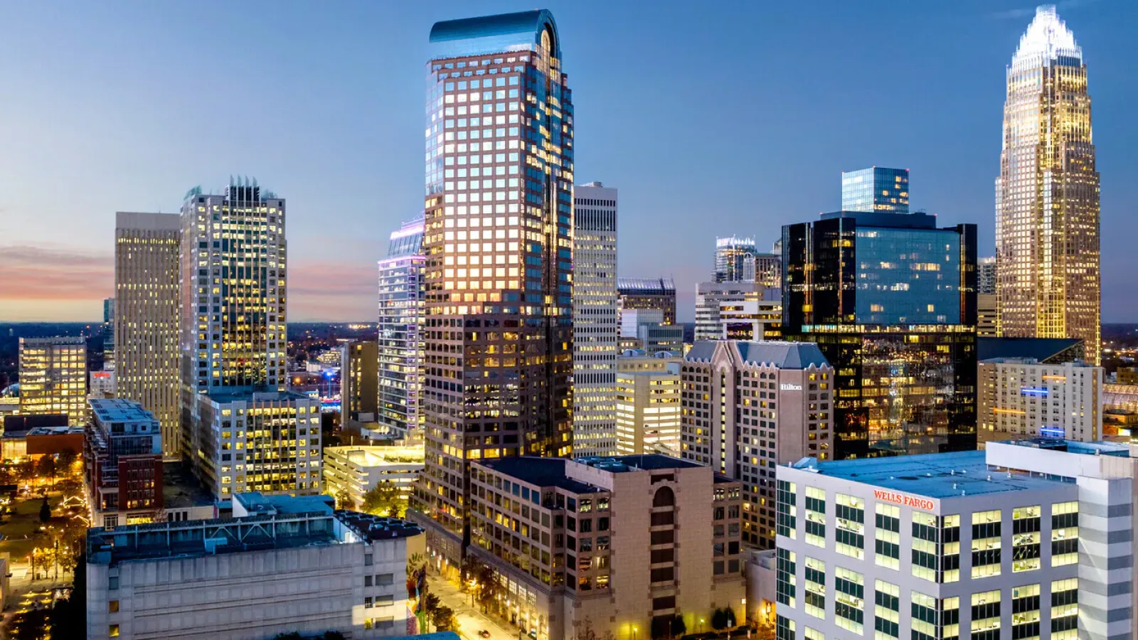 Evening aerial view of a city skyline with illuminated skyscrapers and clear blue sky.