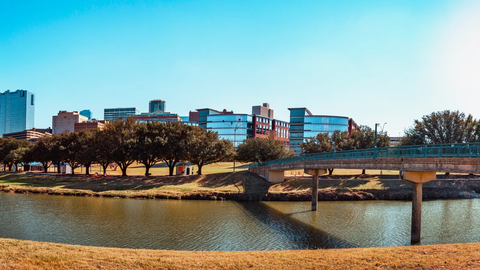 Fort Worth City skyline with modern buildings, trees, river, and pedestrian bridge under a clear blue sky