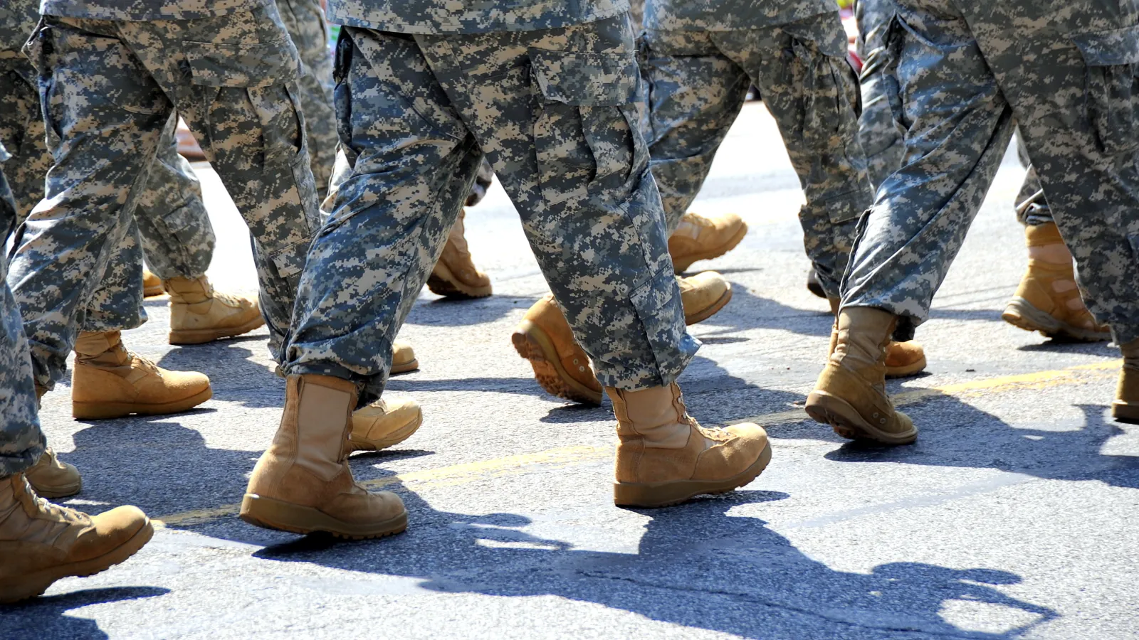 Close-up of soldiers marching in camouflage uniforms and tan combat boots on a sunny day.