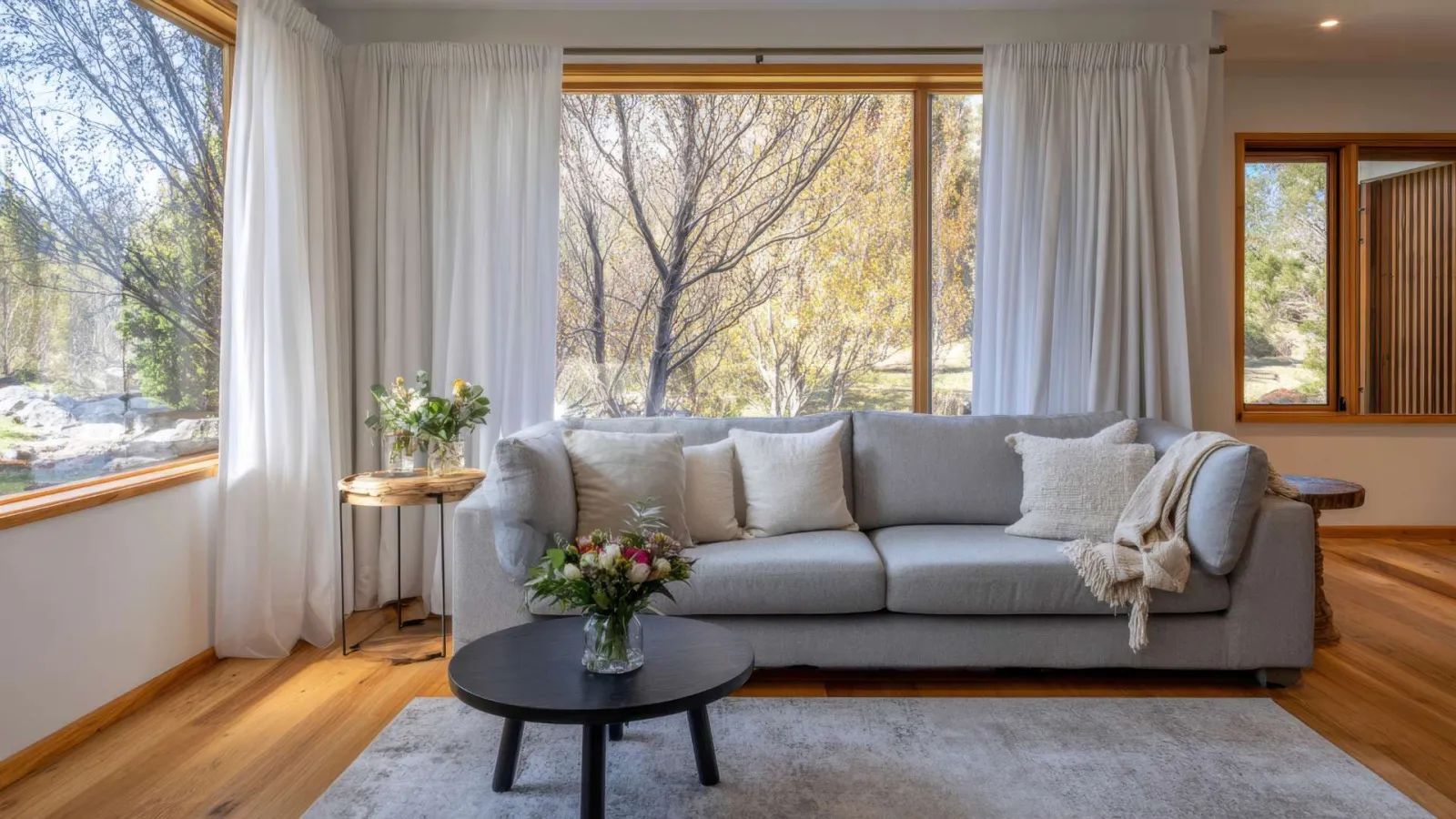 Bright living room with gray sofa, white curtains, wooden floor, round coffee table, and large windows showing trees.