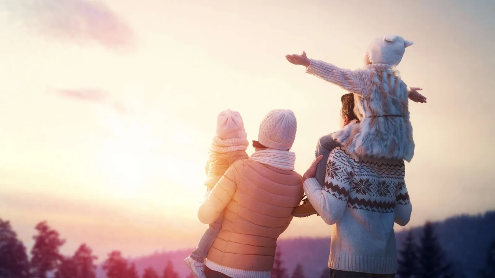 Family of four in winter clothes enjoying a snowy sunset in a forested mountain area.