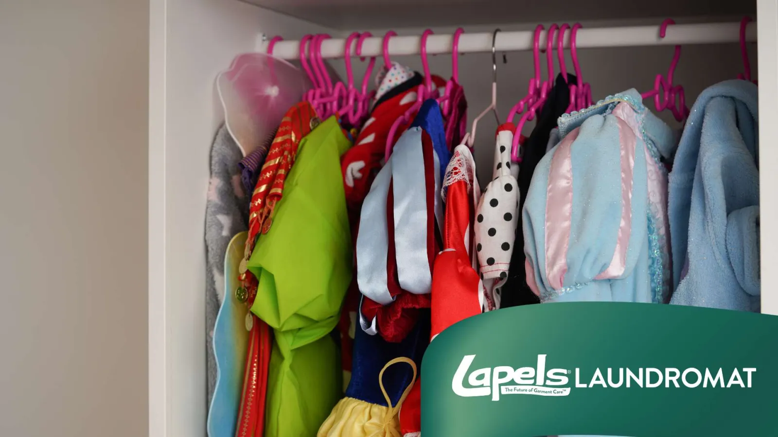 Colorful children's costumes hanging on pink hangers in a closet with Lapels Laundromat branding.