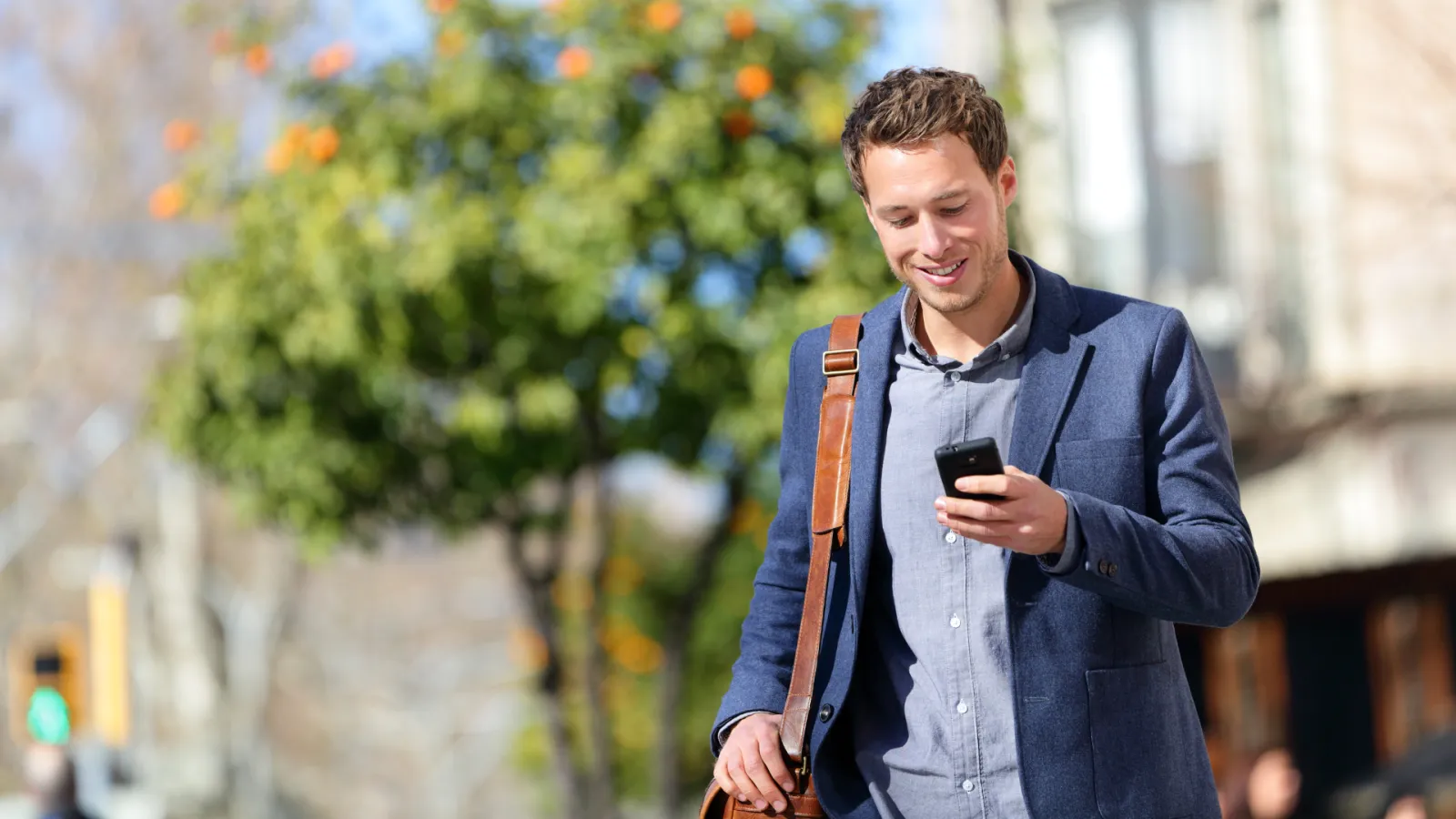 Smiling young man in blue blazer walking outdoors while using smartphone on a sunny day.