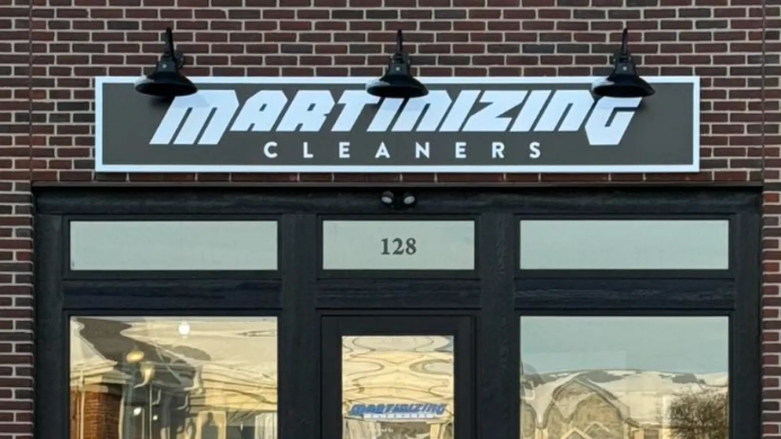 Martinizing Cleaners storefront with black framed glass door, brick wall, and illuminated sign above showing name.