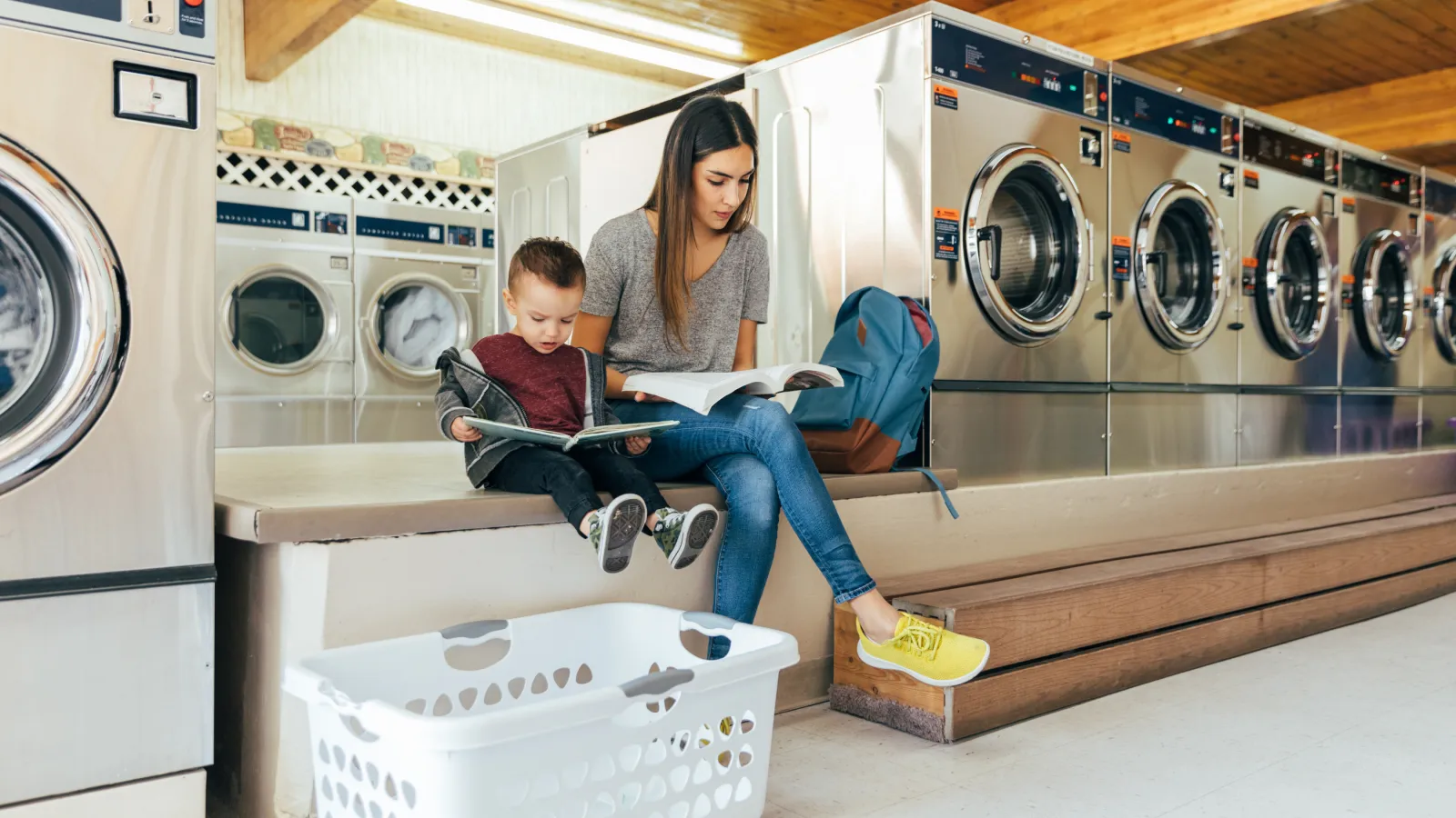Mother and child sitting on bench reading books in laundromat with washing machines and laundry basket nearby