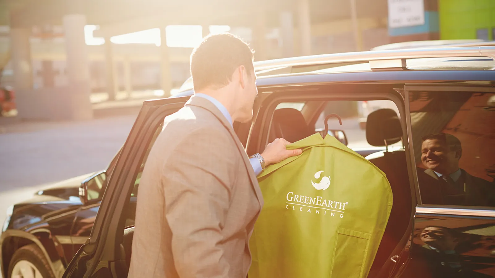 Man in suit holding GreenEarth Cleaning garment bag near open car door in bright sunlight.