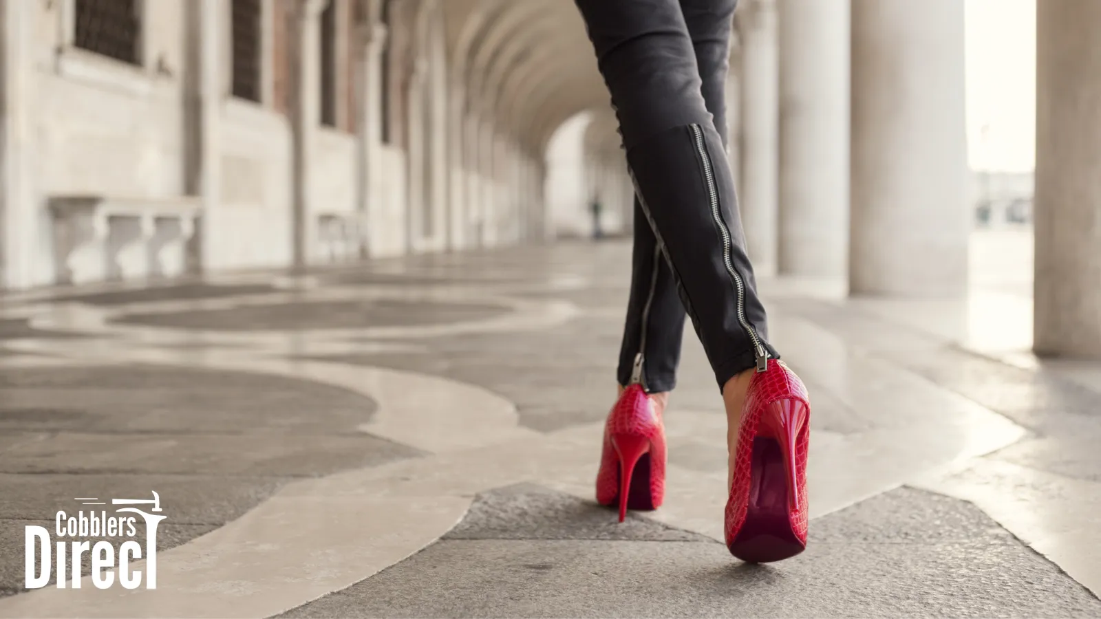 Person walking on stone pavement wearing black pants with zippers and red high heels under an arched colonnade.