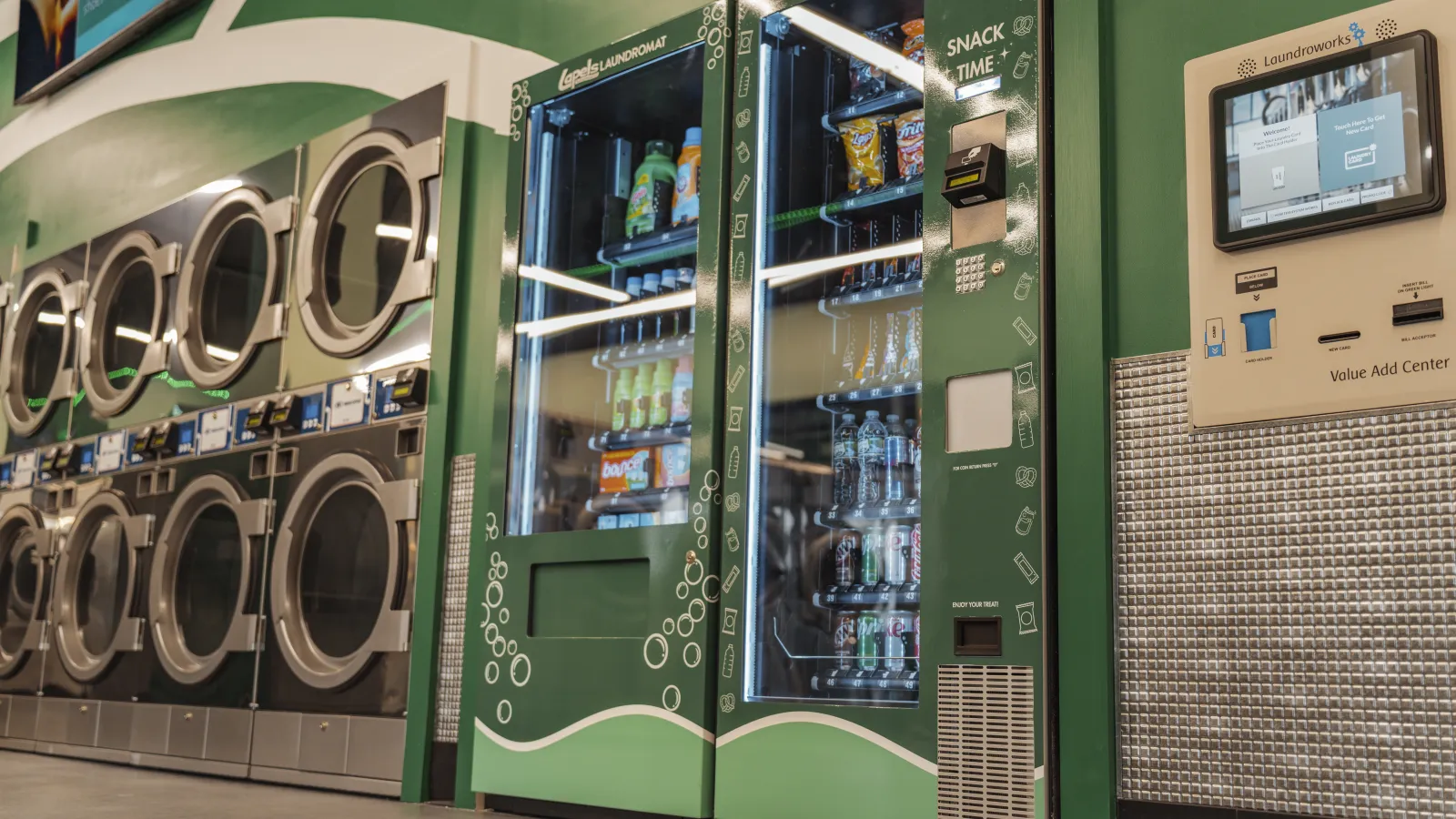 Green snack and drink vending machines next to stainless steel laundromat washers with digital payment kiosk.