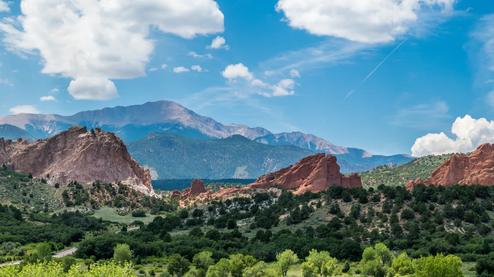 Scenic view of red rock formations and green forest under blue sky with clouds and distant mountains.