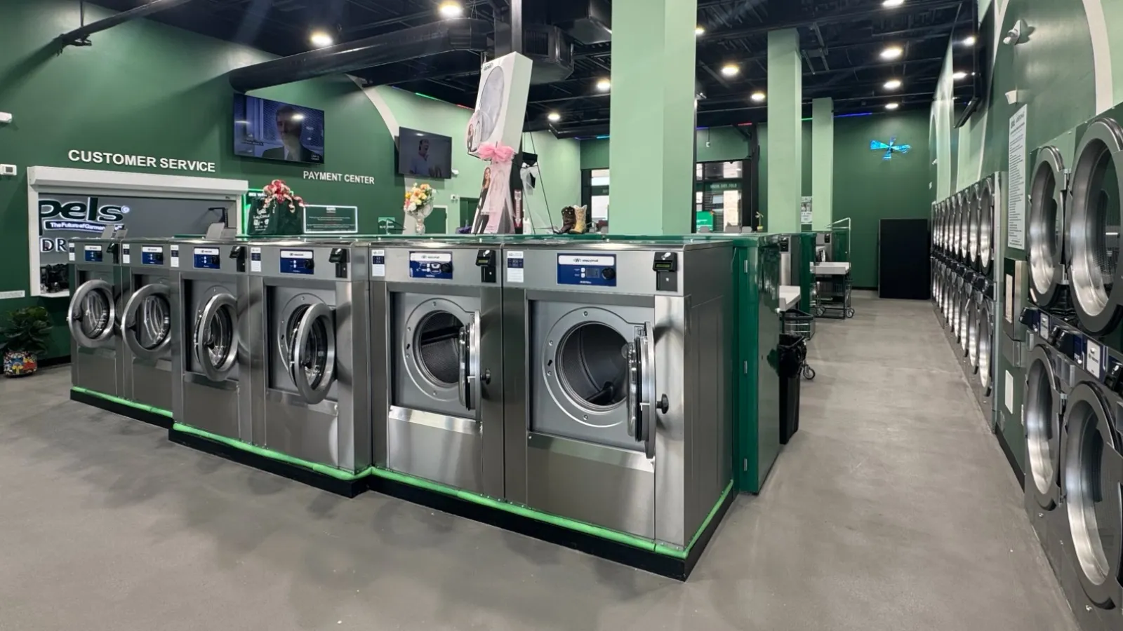 Modern laundromat interior with rows of stainless steel washers and dryers in a clean, green-themed space.