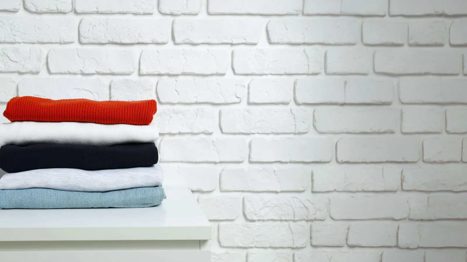 Stack of folded sweaters in red, white, black, grey, and blue on a white surface against a white brick wall.